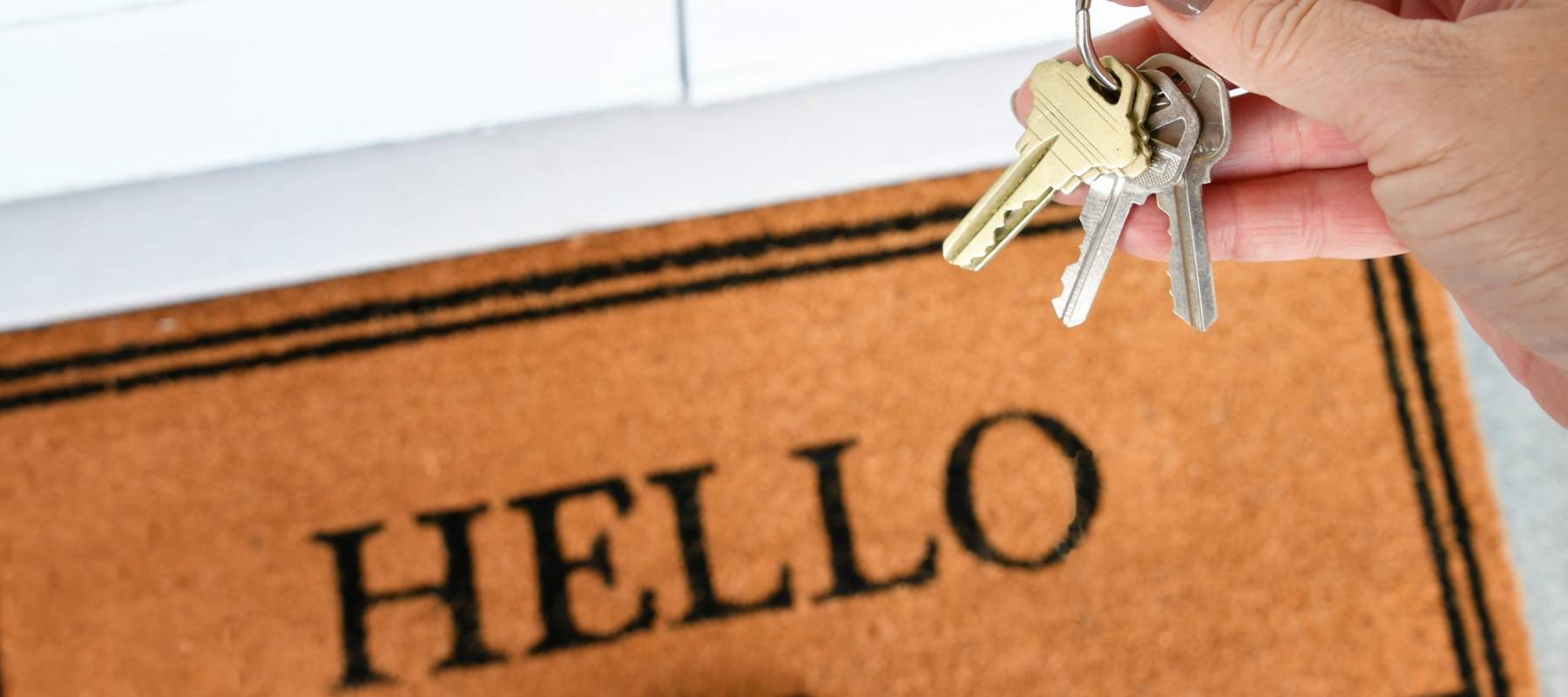 Female hand holding a set of keys over a welcome mat saying Hello - new home