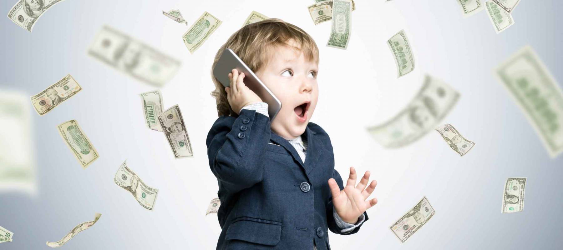 Cute toddler wearing a suit and talking on his smartphone is standing under a dollar rain in a gray room
