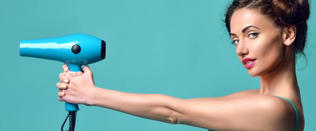 Happy young curly brunette woman with hair dryer on blue mint background