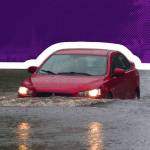 Red car drives through deep flood waters on residential street.