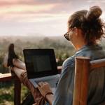 A woman works remotely on her laptop on a porch overlooking a sunset on a beautiful landscape of trees