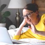 Woman in yellow sweater laying on bed using laptop computer for video chat.