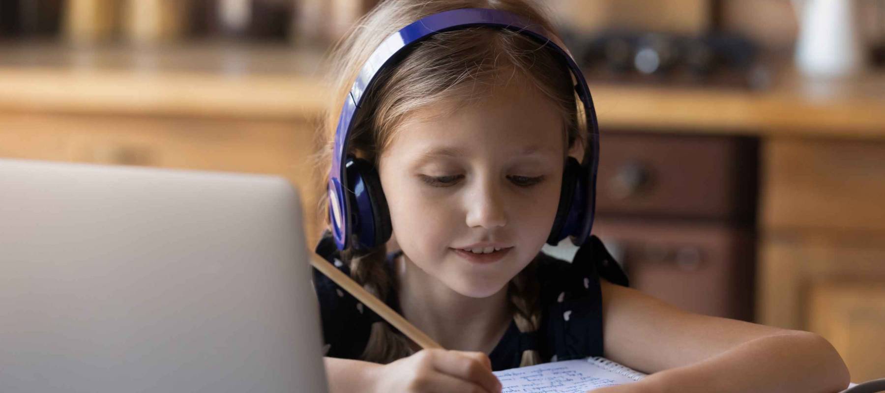 Cute primary schoolkid doing school task at home, listening to audio tutorial, writing in notebook.