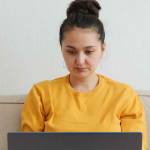 Woman blogger influencer working on the computer at home on the couch. Young asian woman in yellow sweater with focused gaze working remotely at home during pandemic