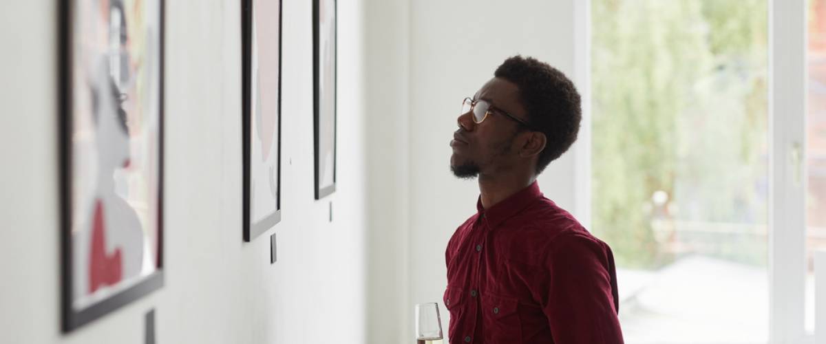 A man views framed artwork at a gallery holding a flute of champagne