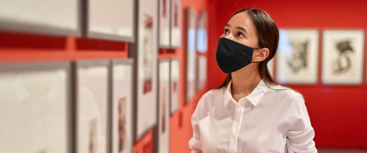 A young woman wearing a mask and holding sunglasses looks at framed photos in an art gallery