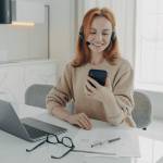 Smiling female office worker in headset using smartphone while sitting at her workplace with laptop