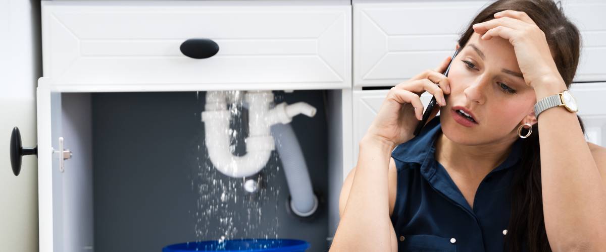 Woman on the phone with a plumber, sitting in front of an open cabinet with a burst pipe dripping into a bucket.