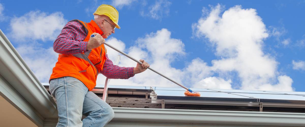 Man in yellow hard hat and orange work vest on a ladder, using a broom to clean a roof.