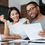Photo of cheerful loving young couple using laptop and analyzing their finances with documents. Look at papers.