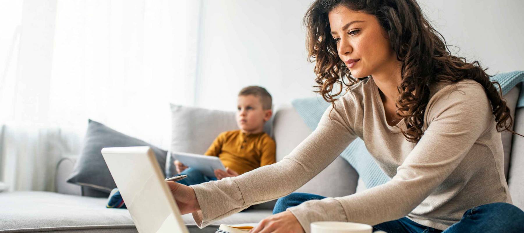 Smiling mom working at home with her child on the sofa while writing an email. Young woman working from home, while in quarantine isolation during the Covid-19 health crisis