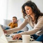 Smiling mom working at home with her child on the sofa while writing an email. Young woman working from home, while in quarantine isolation during the Covid-19 health crisis