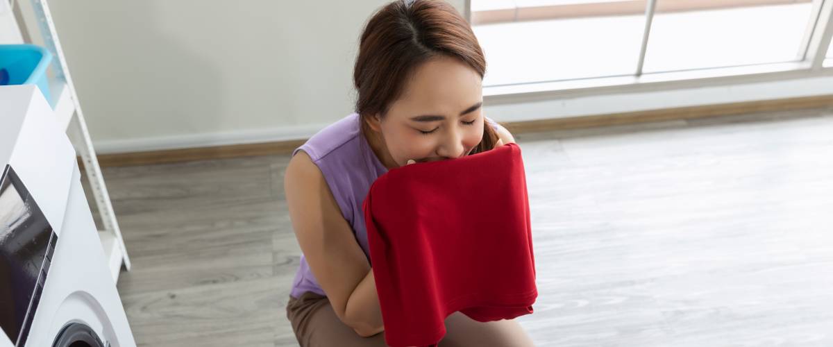 woman kneeling down with a basket of laundry, smelling a red towel