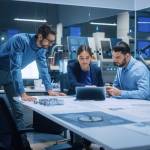 Factory Office Meeting Room: Team of Engineers Gather Around Conference Table, They Discuss Project Blueprints, Inspect Mechanism, Find Solutions, Use Laptop. Industrial Technology Factory