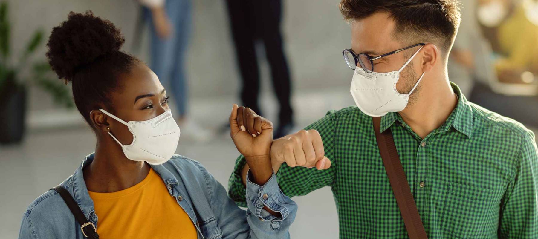 Happy university students greeting in a lobby and elbow bumping while wearing protective face masks.