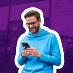 Happy young man sits on a bench outside, looking down at his phone.