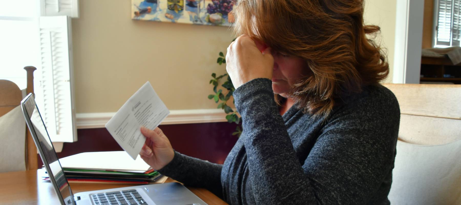 Woman sitting at dining table holding her head in her hands and looking at bills.