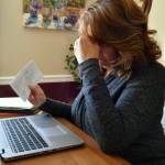 Woman sitting at dining table holding her head in her hands and looking at bills.
