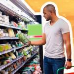 Man stands in produce aisle at grocery store holding a basket and looking at a box of greens.