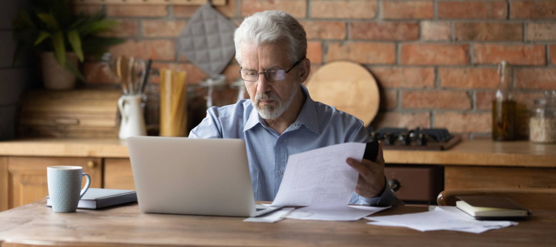 Older man sitting at desk, looking at files quizzically.