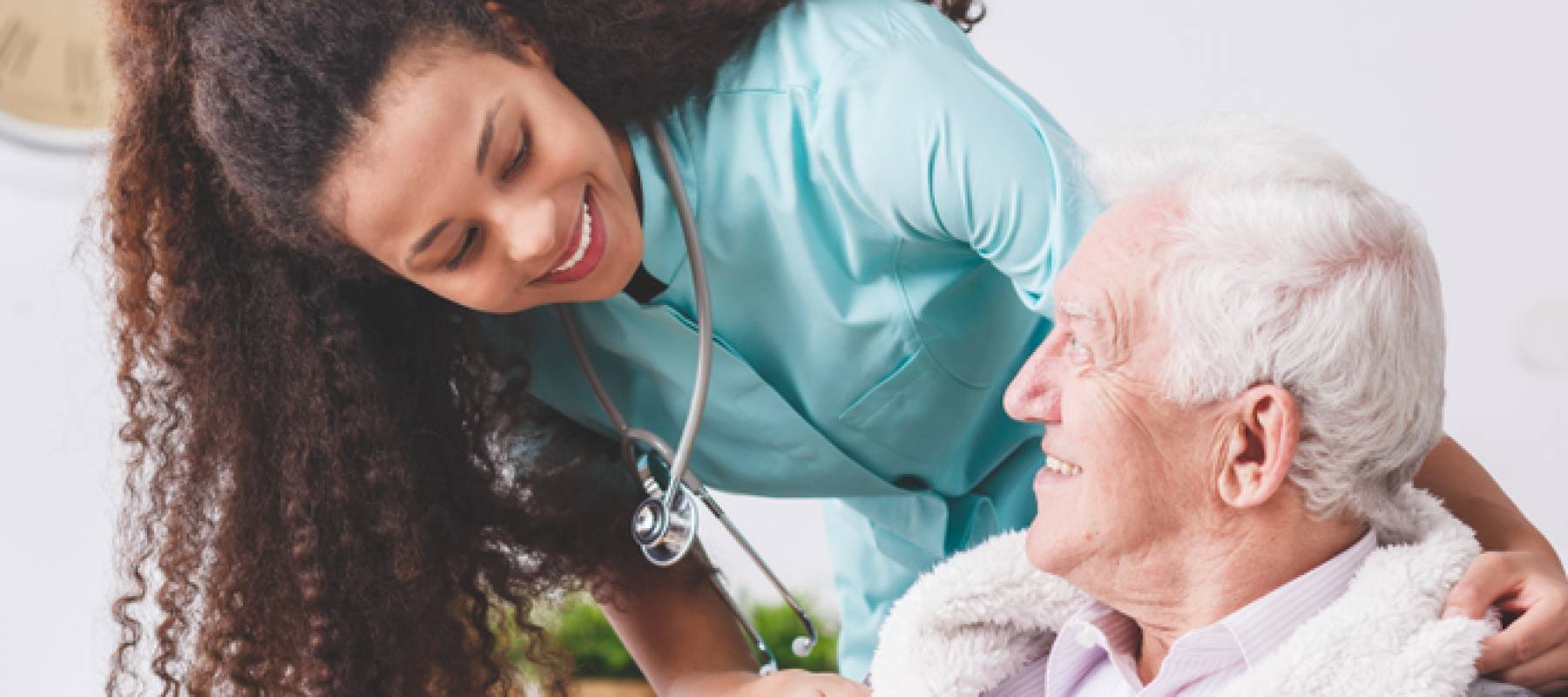Smiling nurse leans over an older man in a wheelchair, smiling up at her.