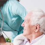 Smiling nurse leans over an older man in a wheelchair, smiling up at her.