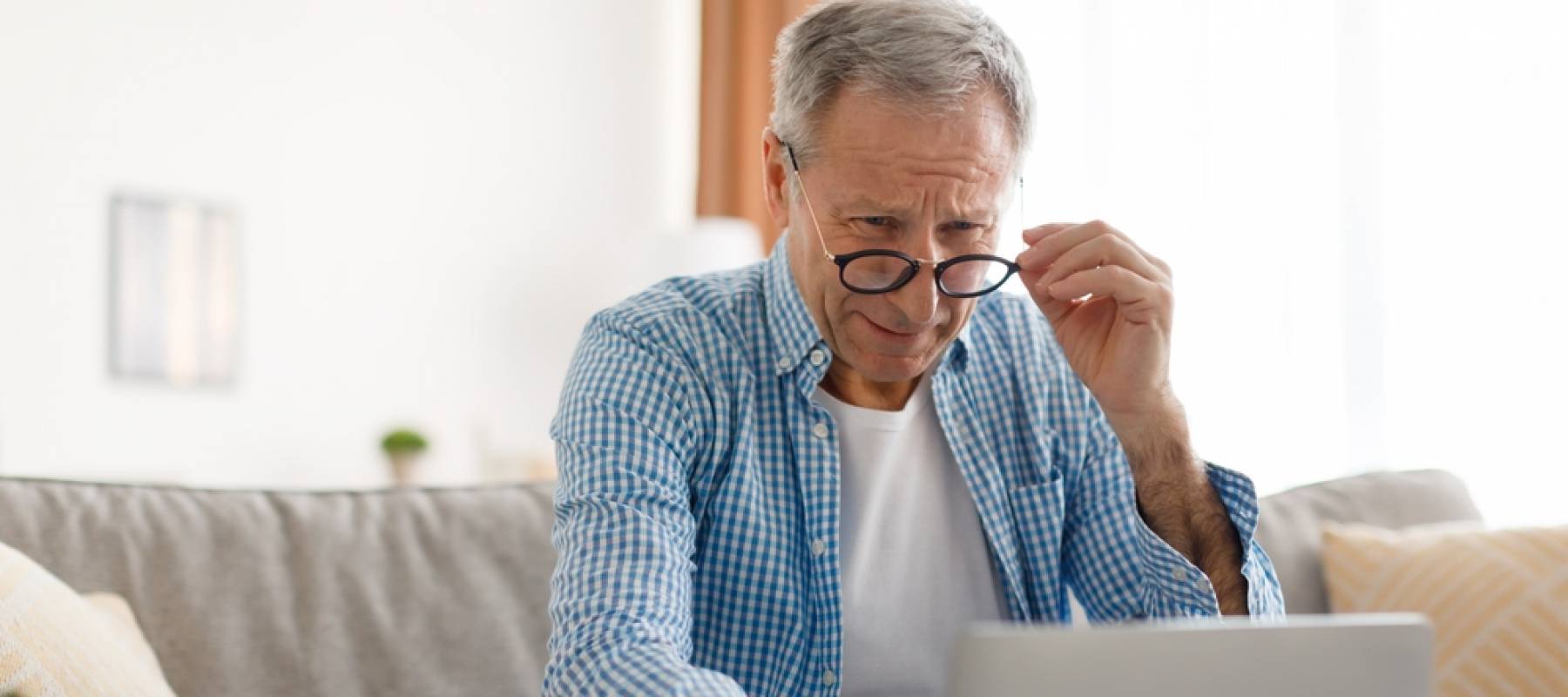 Older man sits on couch looking at computer with puzzled look on his face.
