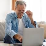 Older man sits on couch looking at computer with puzzled look on his face.