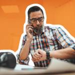 Man talking on the phone and looking at his watch, while sitting at a desk.