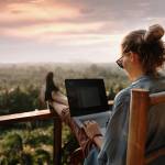 A woman using her computer with her feet up on a deck rail overlooking a wooded vista