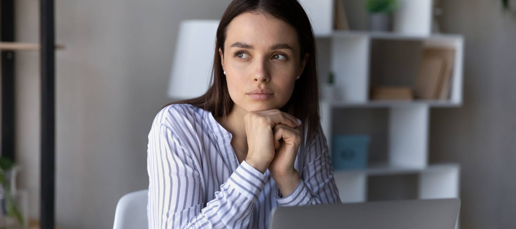 Serious thoughtful employee sitting at table with laptop, looking at window away, thinking over business problems