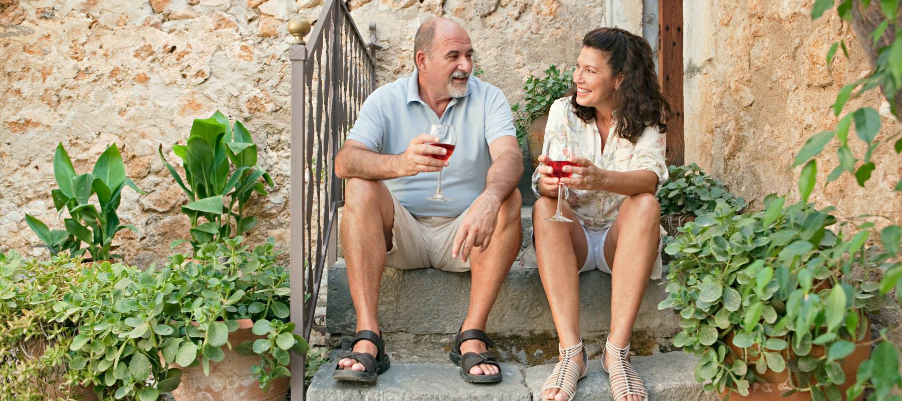 Attractive senior couple sitting on the steps of a luxury holiday villa with stone walls during a summer holiday, drinking wine and having a conversation.