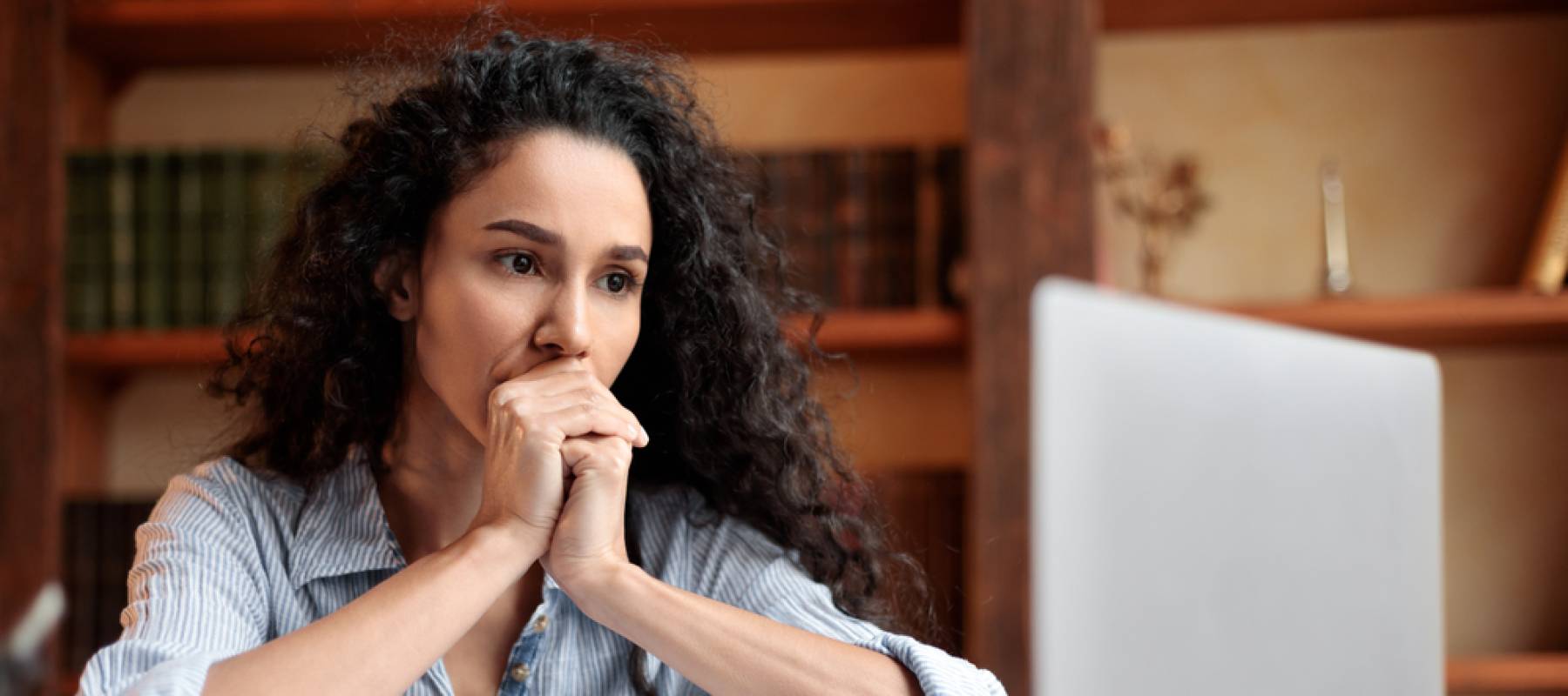 Woman looks at her computer with hands clasped on her chin, looking concerned.