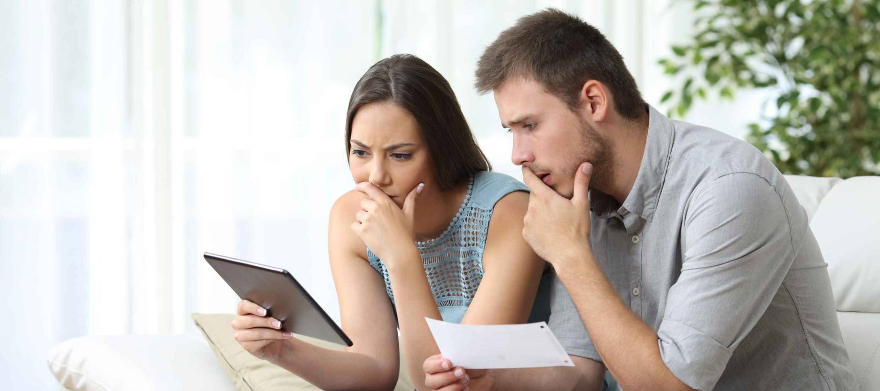 Worried couple comparing receipt on tablet sitting in the sofa in the living room at home