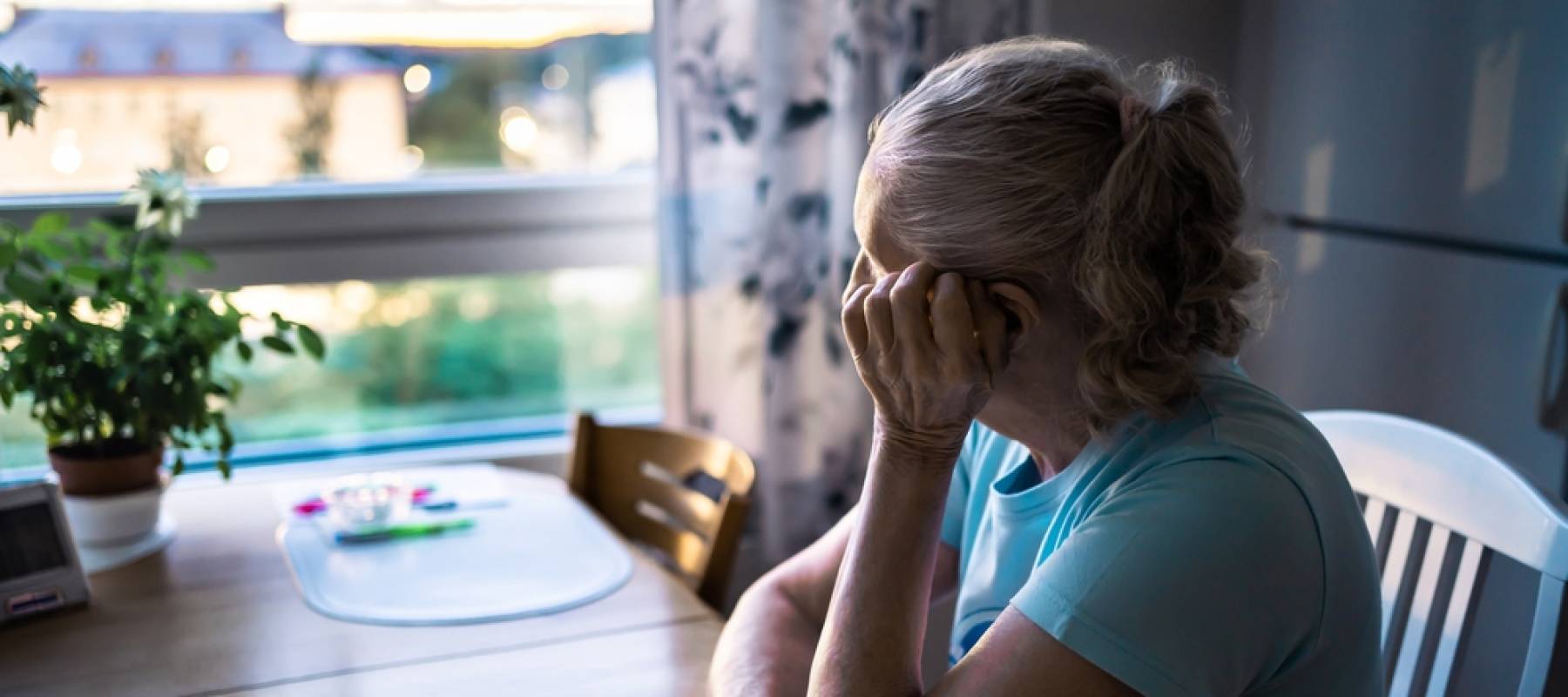 Woman turned away from the camera, sitting at a kitchen table and looking out a window.