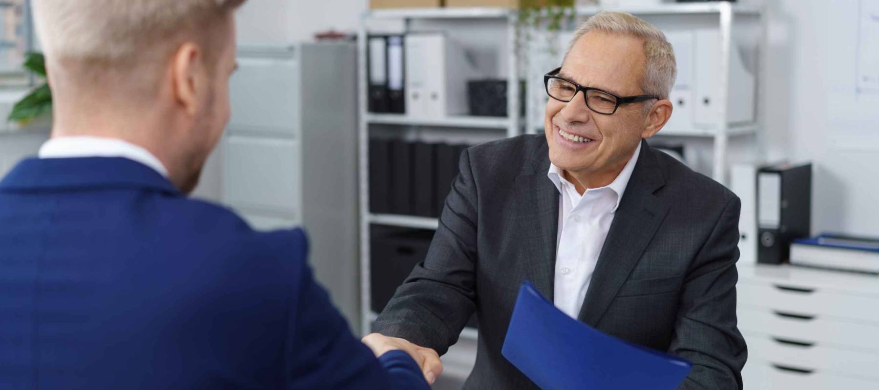 Middle aged boss in eyeglasses and dark suit shaking hands with young job seeker in blue