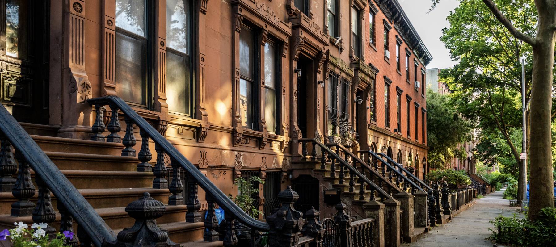 Scenic view of a classic Brooklyn brownstone block with a long facade and ornate stoop balustrades on a summer day in Clinton Hills, Brooklyn