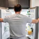 Back of hungry man opening fridge refrigerator doors domestic appliance searching for food inside with condiments and juice in modern kitchen