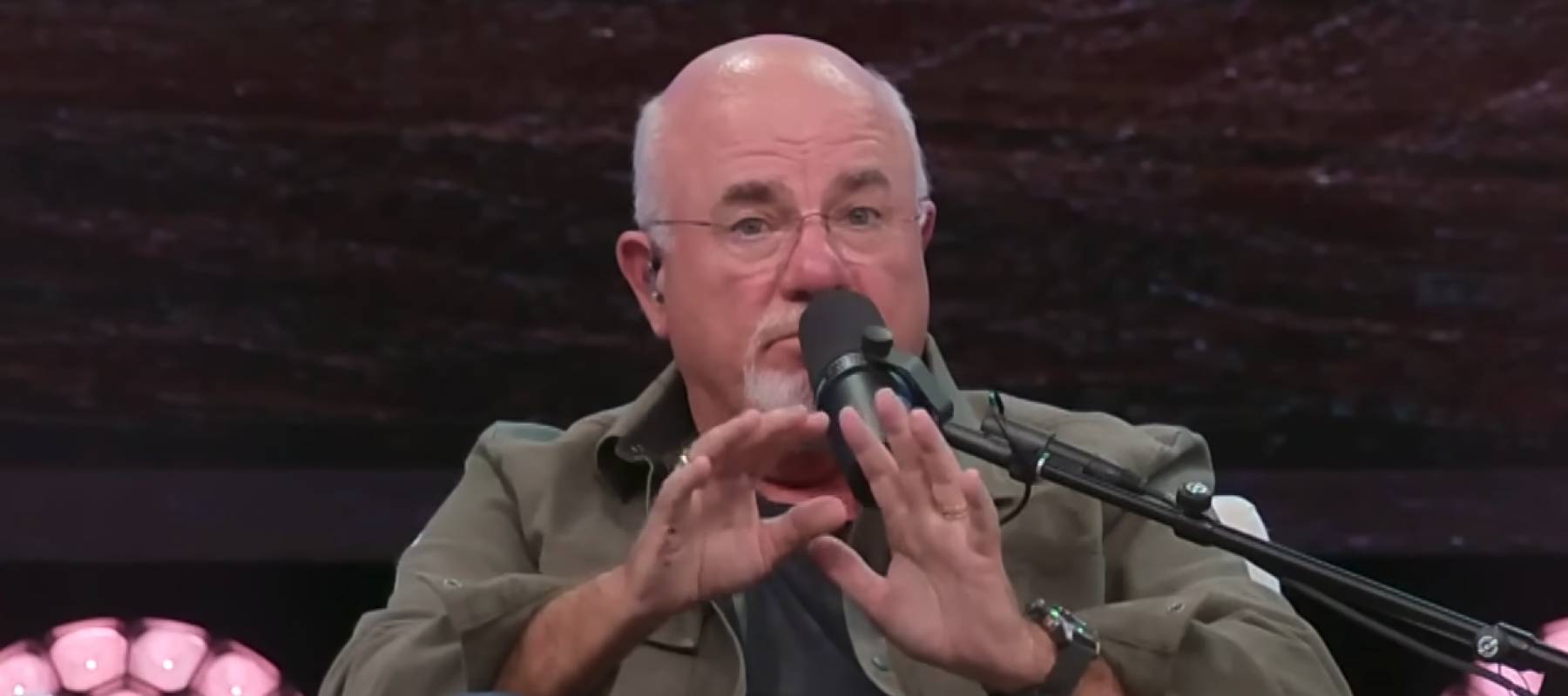 Dave Ramsey sits in a chair on a stage, talking into a microphone and making a surprised face.