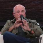 Dave Ramsey sits in a chair on a stage, talking into a microphone and making a surprised face.