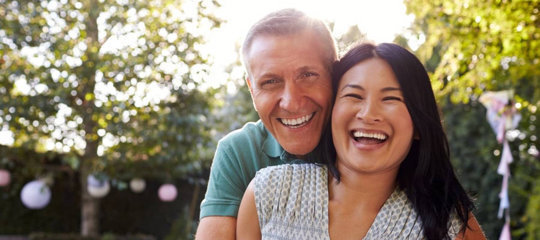 Mature couple smile and laugh posing for the camera with man's arms draped around woman from behind.