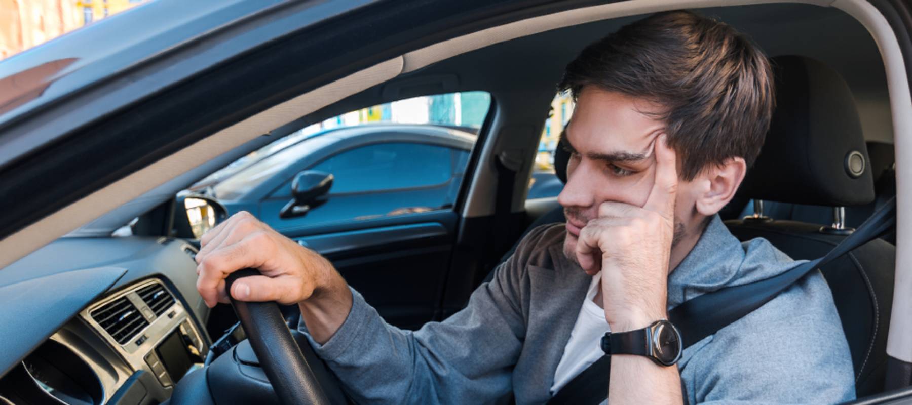 Annoyed tired-looking young man sitting in driver&#039;s seat of car with arm on the wheel.