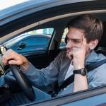 Annoyed tired-looking young man sitting in driver's seat of car with arm on the wheel.