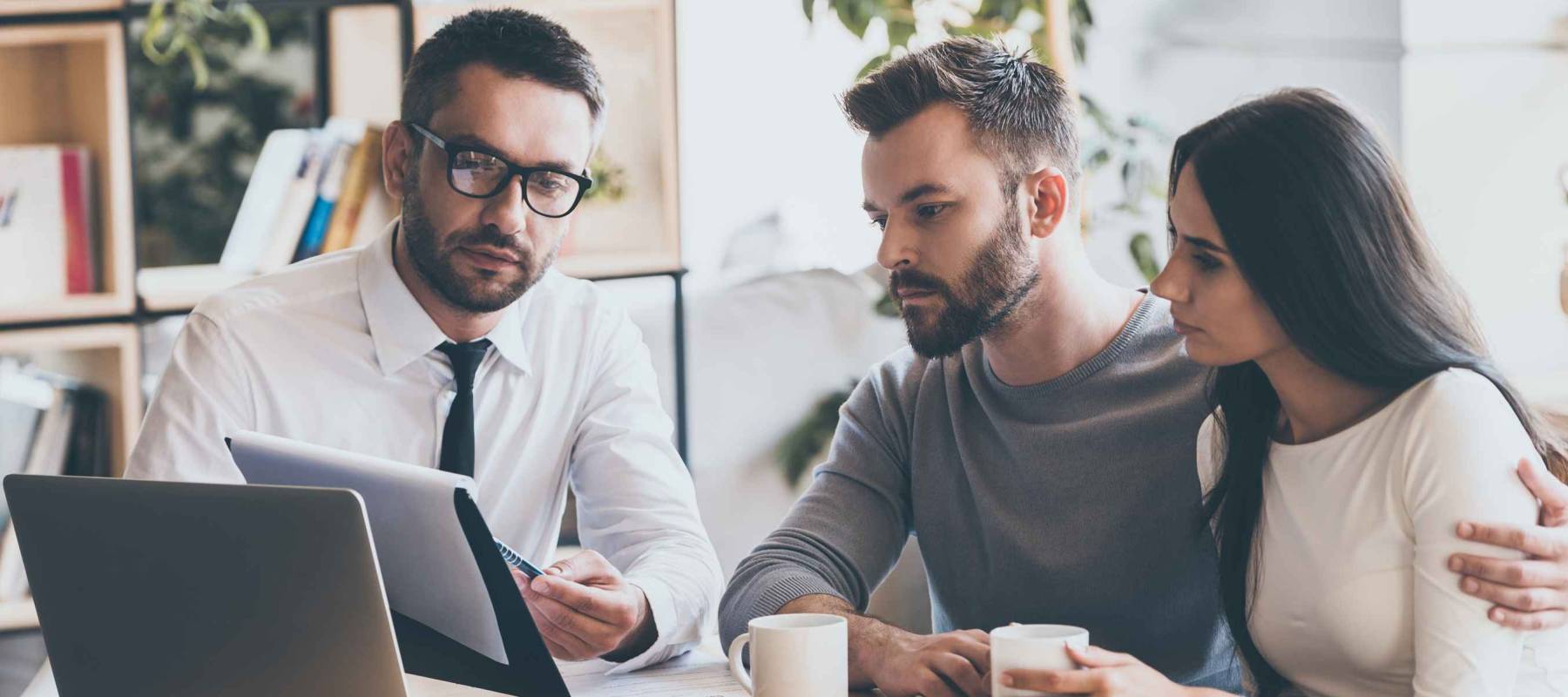 I need your signature here. Confident young man in shirt and tie holding some document and pointing it while sitting together with young couple at the desk in office