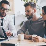 I need your signature here. Confident young man in shirt and tie holding some document and pointing it while sitting together with young couple at the desk in office