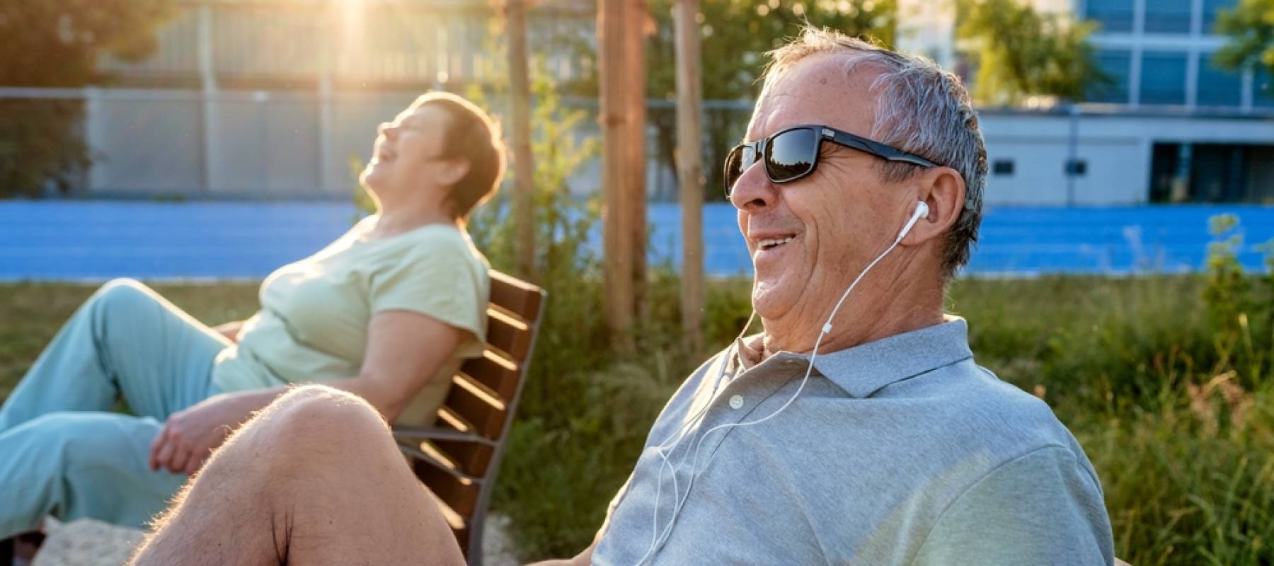Happy smiling senior couple with earphones exercising using outdoor training exercise bike machine at municipal sports ground on bright sunny summer day.