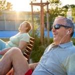 Happy smiling senior couple with earphones exercising using outdoor training exercise bike machine at municipal sports ground on bright sunny summer day.