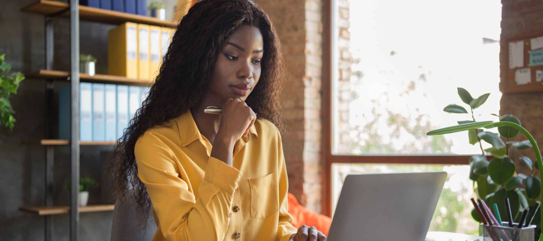 Photo of thoughtful dark skin woman browse internet laptop on table think business plan wear yellow shirt indoors in office