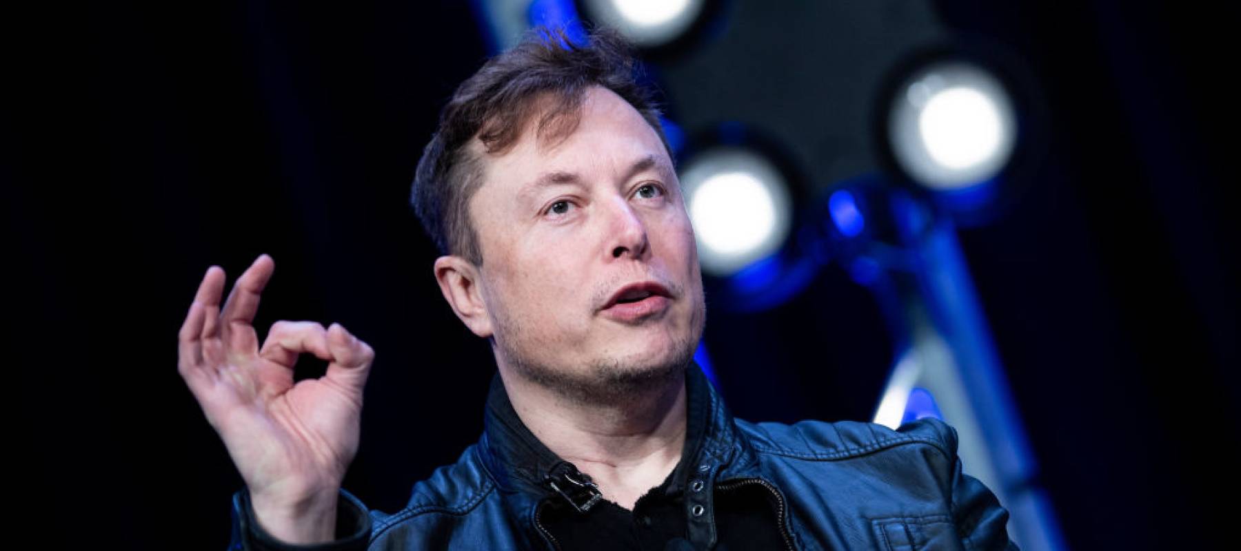Elon Musk speaks on stage with a black background, holding up his hands.
