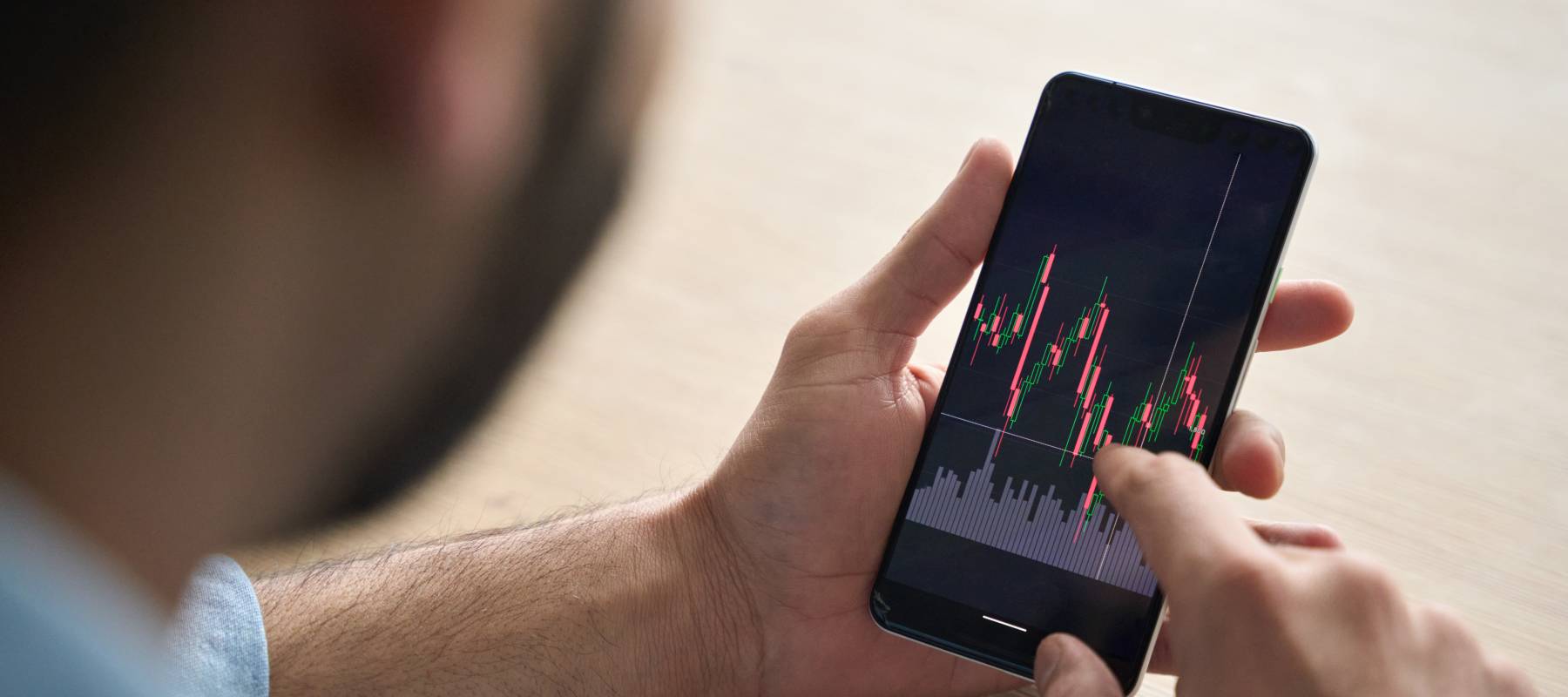 Over shoulder close up view of male analyst broker's hands holding touch screen device smartphone using tech ecommerce application of finance stock markets with graphs and numbers.